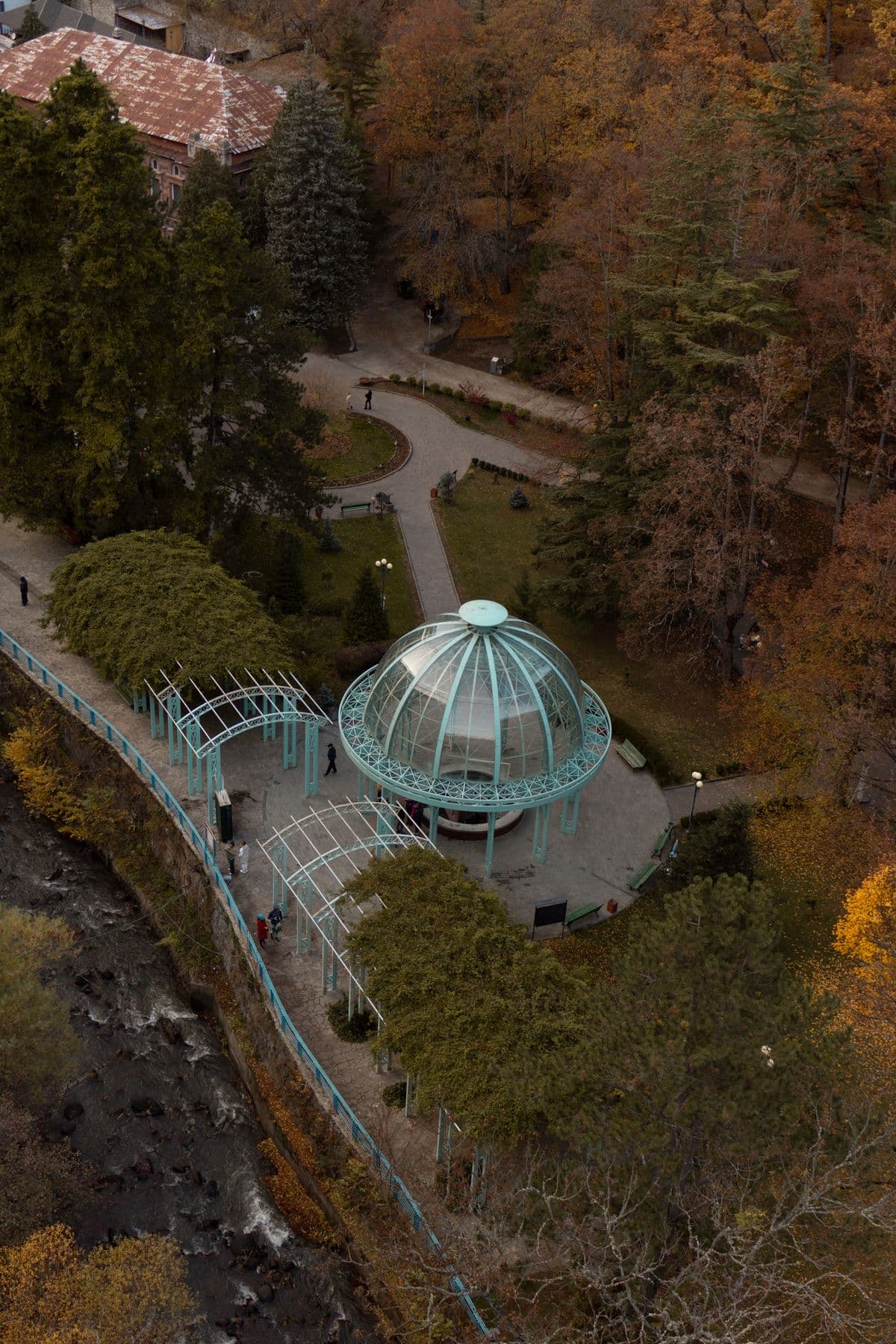 An elevated view of the green glass-domed spring pavilion in Borjomi's park, ringed by paths and benches among autumn trees, with the rocky river below