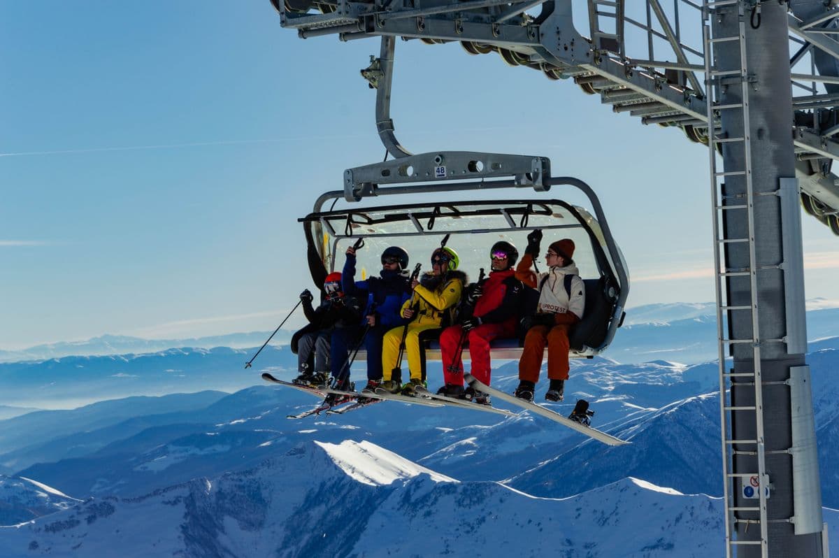 Five riders on a Gudauri chairlift with skis and a snowboard hanging below, hazy blue ridges far beneath them