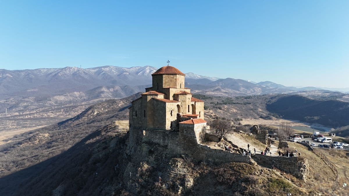 The stone church of Jvari with its red-tiled roofs on a rocky hilltop, bare hills and snow-dusted mountains behind