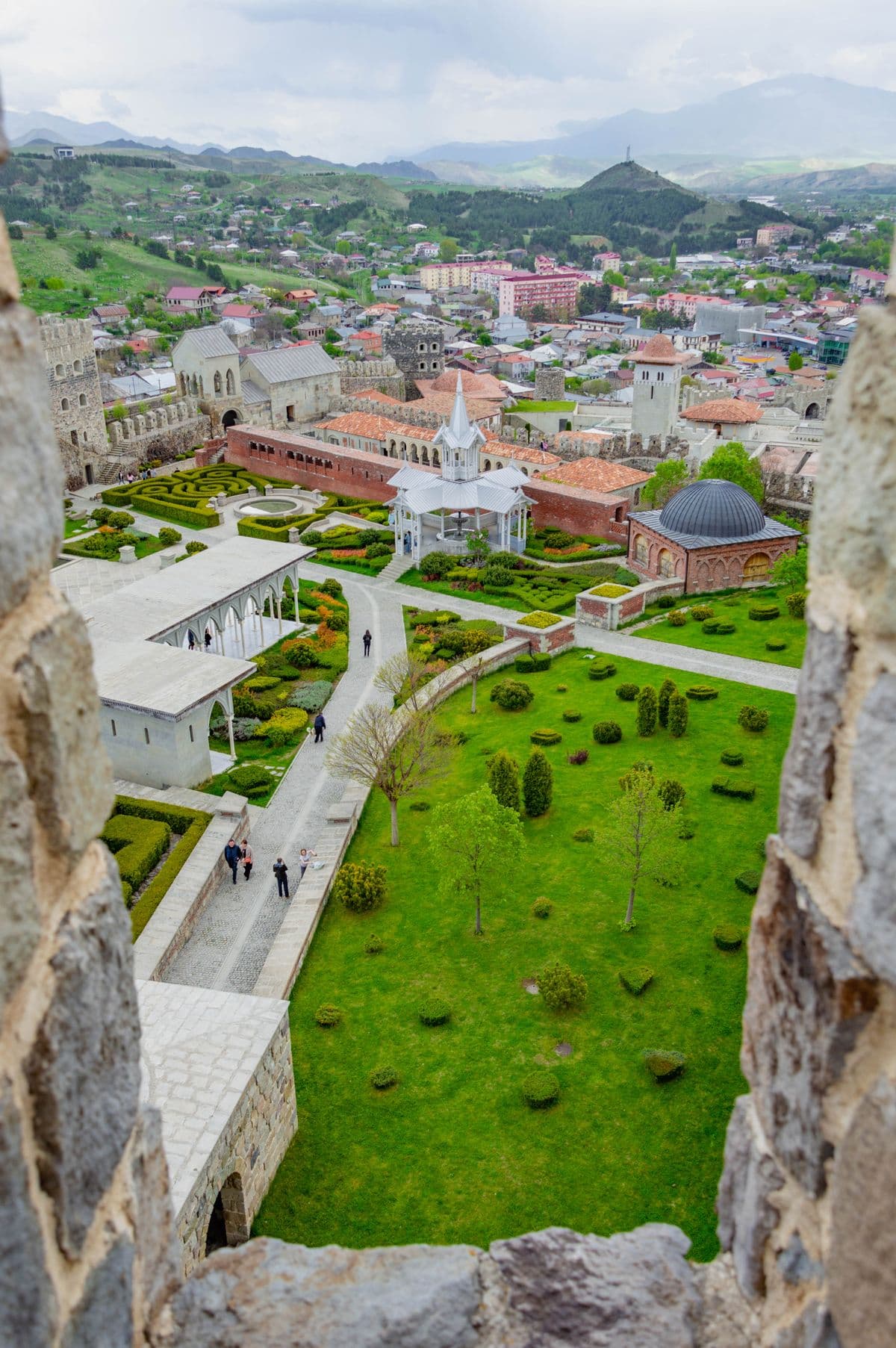 Looking down through a gap in Rabati's stone battlements onto the fortress gardens, a white pavilion and a domed brick building, with Akhaltsikhe and green hills behind