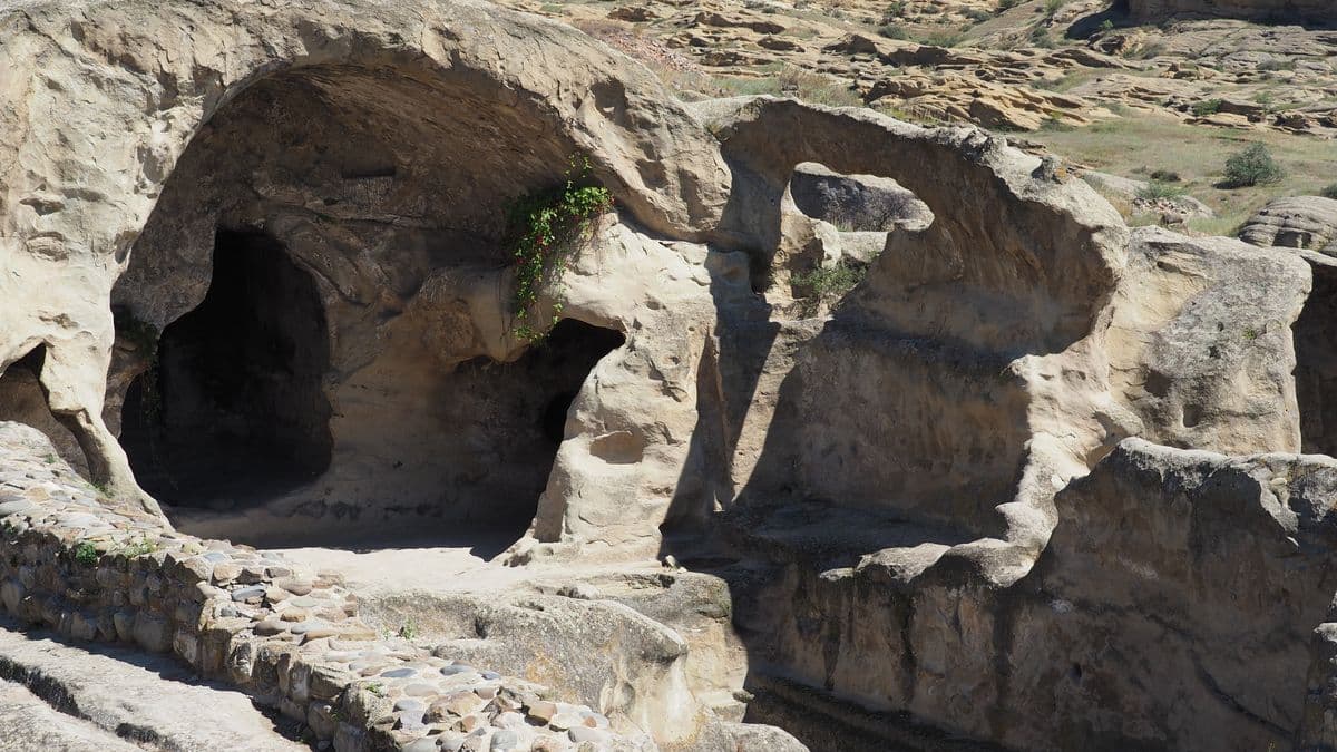 Pale sandstone chambers cut into the rock at Uplistsikhe, one with a carved vaulted ceiling, a cobbled path running past