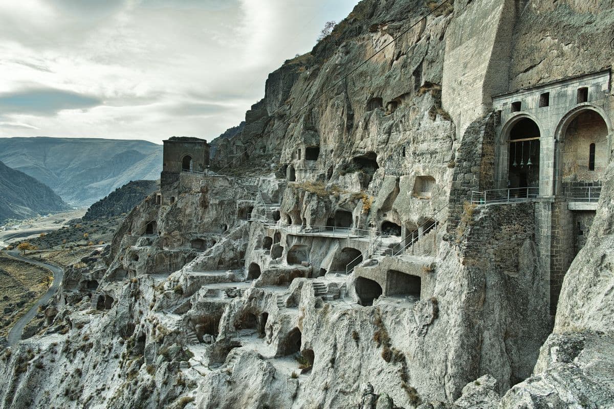 The cave city of Vardzia: tiers of rock-cut chambers, stairways and railed walkways on a cliff face, with an arched stone bell tower on the right and a valley road below